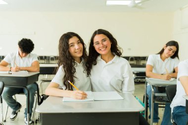 Üniforma giyen iki mutlu lise öğrencisi sınıfta birlikte ders çalışırken aynı anda gülümsüyor.