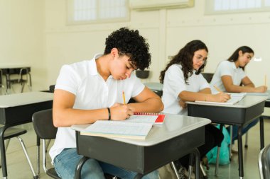 Sınıfta sıralarında otururken not defterlerine yazan bir grup odaklanmış Latin lise öğrencisi.