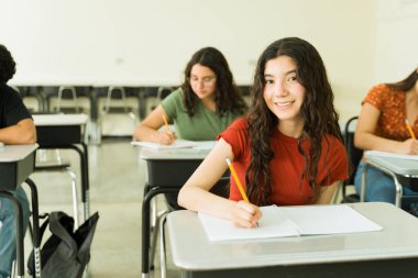 Gülümseyen kız öğrenci, bir lise dersinde defterine özenle yazarak öğrenme süreciyle meşgul.