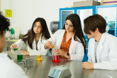 Laboratuvar önlüğü ve güvenlik gözlüğü takan genç kimya öğrencileri bilim laboratuvarında fen dersi sırasında renkli kimyasalları karıştırarak deney yapıyorlar.