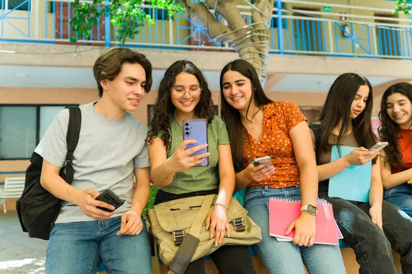 Bir grup neşeli Latin lise öğrencisi ders aralarında akıllı telefonlarını kullanıyor, sosyal medya içeriğini paylaşıyor ve tatillerinin tadını çıkarıyorlar.