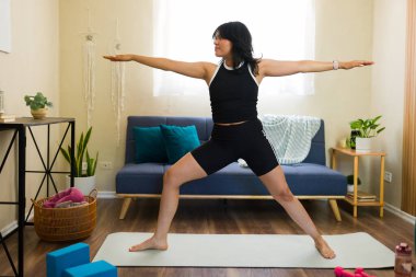 Evde yoga yapan genç bir kadın. Oturma odasındaki paspasın üzerinde iki poz vererek sağlığını ve sağlığını yükseltiyor.