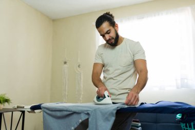 İspanyol bir adam düzenli yatak odasındaki kıyafetleri ütüleyerek ev işlerini yapıyor. Ev hayatının ve organizasyonun rutinini yansıtıyor.