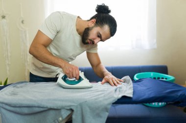 Ütü masasında gömlek ütüleyen, ev işlerini rahat ve davetkar bir ortamda yapan bir adam.