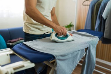 Genç bir adam ütü masasında gömlek ütülüyor, rahat bir ev ortamında ev işi ve günlük ev işleri yapıyor.