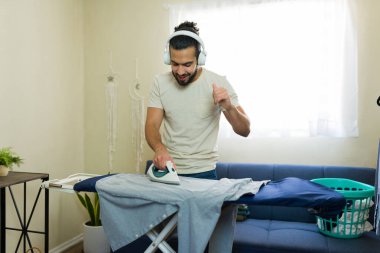 Latin adam ütü masasında giysileri ütülüyor ve kulaklıkla müzik dinliyor, ev işlerinin keyfini çıkarıyor.