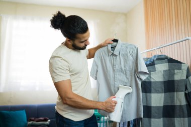 Askıda asılı olan gömleğin kırışıklıklarını silmek için taşınabilir buharlı gemi kullanan İspanyol bir adam. Ev işlerini idare ediyor ve düzeni sağlıyor.