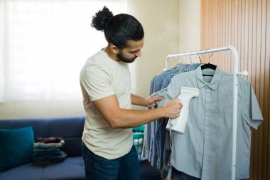 Genç bir adam taşınabilir bir buhar makinesi kullanarak evde giysilerini buharlaştırıyor, gömlekleriyle ilgileniyor ve ev işi yapıyor.