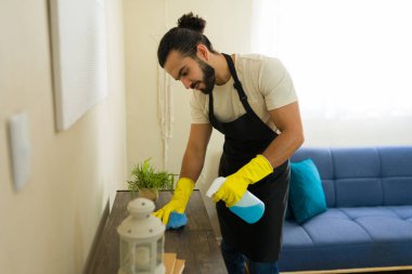 Önlük ve eldiven giyen genç adam oturma odasındaki mobilyaları dezenfektan sprey ve bezle temizliyor.