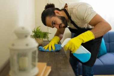 Önlük ve sarı eldivenler giyen genç erkek hademe mavi dezenfektan spreyi ve bez parçasıyla ahşap bir yüzeyi temizliyor.