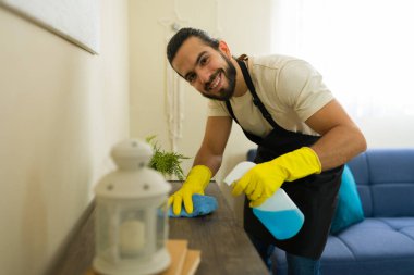 Mutlu profesyonel temizlikçi önlük ve sarı eldivenlerle ahşap mobilyalara dezenfektan sıkar ve ev işi yaparken mavi bez ile silerdi.
