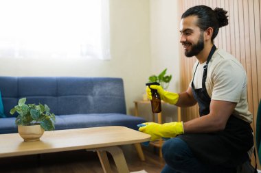 Önlük ve eldiven giyen genç bir adam oturma odasında kahve masasını temizliyor, yüzeye dezenfektan sıkıyor.