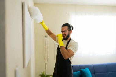 Önlüklü ve eldivenli genç bir adam toz beziyle duvarı temizliyor, alerji belirtileri gösteriyor ve burnunu kapatıyor.