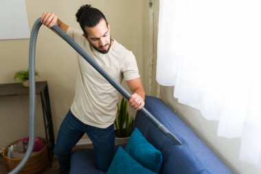 Adam oturma odasında mavi bir kanepeyi süpürüyor, ev işlerini yapıyor ve ev ortamını temiz tutuyor.