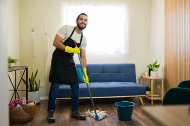 Önlük ve eldiven giymiş gülümseyen erkek hademe rahat bir oturma odasında yerleri paspaslıyor, düzenli ve lekesiz bir ev ortamı için profesyonel temizlik hizmeti veriyor.