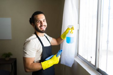 Önlük giymiş, sarı eldivenli, gülümseyen bir erkek hademe elinde mavi bir bez tutarken cam camına temizlik malzemesi sıkıyor.