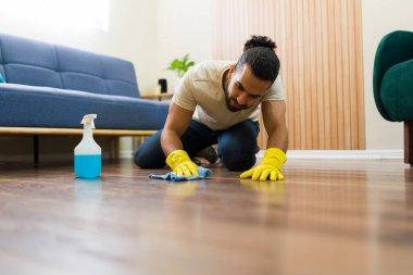 Sarı eldivenler giyen genç adam evinin ahşap zeminini deterjan ve mikrofiber bezle temizliyor, ev işi yapıyor.