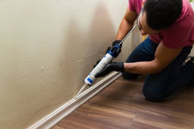 Siyah eldivenli tamirci süpürgelik ve duvar arasına silikon dolgu macunu sürüyor, ev onarımı ve diy projesi.