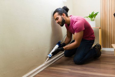 Delikanlı, ev tadilatı sırasında duvar ve etek panosu arasında diz çöküp silikon dolgu macunu sürüyor.