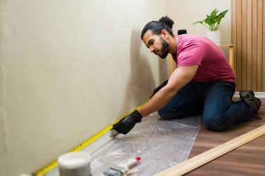Latin müteahhit yerde diz çökerken mezura kullanarak duvarın ölçüsünü alıyor, ev onarımı ve yenilenmesi için hazırlanıyor.