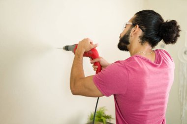 Güvenlik gözlüğü takan, elektrikli matkap kullanan, ev yenileme projesi sırasında duvarda delik açan bir tamircinin arka görüntüsü.