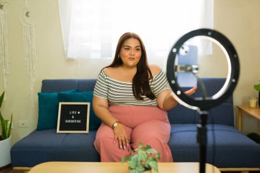 Latin içerik yaratıcısı ev stüdyosundan bir video blogu kaydediyor, optimum ışıklandırma için bir halka ışığı kullanıyor, kanalı abone gibi bir tabela ile tanıtıyor