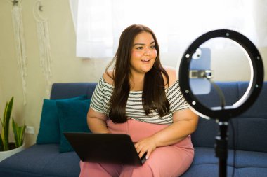 Mutlu büyük ölçekli içerik oluşturucu evde bir vlog kaydediyor bir dizüstü bilgisayar ve akıllı telefonlu bir halka ışığı kullanarak