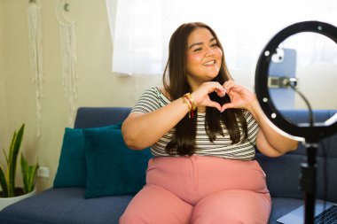 Latince artı boyut etkileyicisi, oturma odasındaki kanepenin rahatlığından video blogu kaydederken elleriyle kalp şekli yaratıyor.