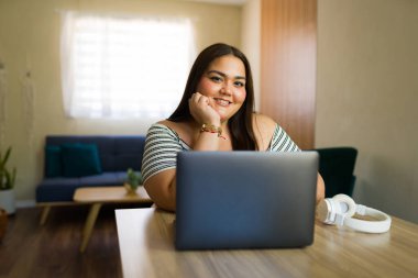 Gülümseyen büyük boyutlu içerik yaratıcısı evde çalışıyor, bilgisayarını meşgul ediyor ve rahat, modern bir oturma odasında kulaklık takıyor.