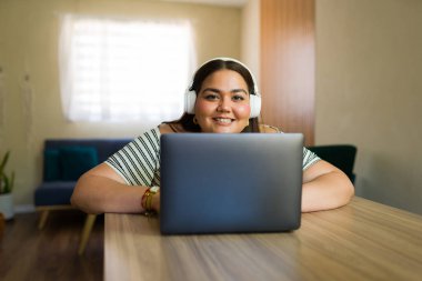 Evde dizüstü bilgisayarla çalışırken kulaklık takan ve gülümseyen iri yarı bir kadın, modern ev hayatı tarzını somutlaştırıyor.