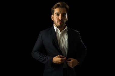 Kendine güvenen işadamı, stüdyoda karanlık bir arkaplanda poz verirken ceketinin düğmelerini ilikliyor.