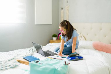 Odaklanmış ergen kız not yazıyor ve ödevlerini yapıyor. Yatak odasında dizüstü bilgisayarını kullanıyor ve okul malzemelerini de yanında taşıyor.