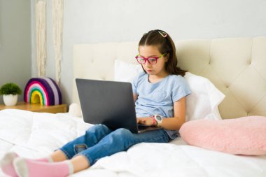 İlkokul öğrencisi gözlük takıyor. İnternette öğrenim, uzaktan eğitim ve evde eğitim için dizüstü bilgisayar kullanıyor. Yatak odasında rahat bir şekilde oturuyor.