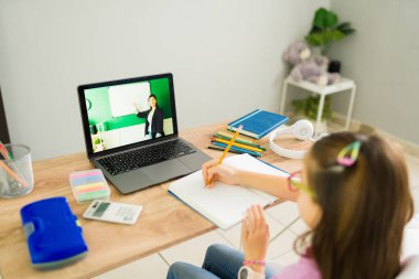 Evde dizüstü bilgisayar kullanan bir bayan öğretmenle çevrimiçi bir sınıfta İngilizce öğrenirken not alan ilkokul öğrencisi kız.