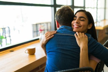 Kız arkadaş kafede erkek arkadaşına sarılıyor, romantik bir buluşmada sevgi ve şefkat göstererek birlikte hassas bir anın tadını çıkarıyor.