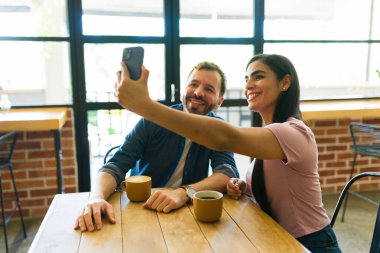 Gülümseyen bir çift akıllı telefonlarıyla neşeli bir selfie çekerken aynı zamanda sıcak bir kafede kahvenin tadını çıkarıyor, unutulmaz bir anı paylaşıyorlar.