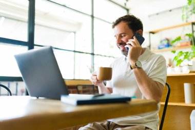 Kafede uzaktan çalışan mutlu erkek serbest çalışan, akıllı telefonla konuşurken bir fincan kahvenin tadını çıkaran ve dizüstü bilgisayar kullanan