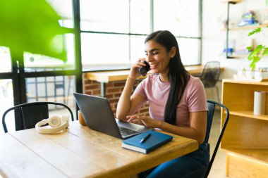 Genç bayan serbest çalışan gülümseyen ve telefonla konuşan, kahve dükkanında dizüstü bilgisayarla çalışan, uzaktan çalışma esnekliğinin keyfini çıkaran