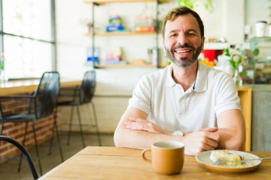 Gülümseyen adam, sıcak bir dekorla çevrili sıcak bir kafede kahve ve pastayla huzurlu bir anın tadını çıkarıyor.