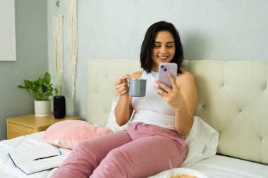 Gülümseyen serbest çalışan bir kadın cep telefonu kullanıyor ve evde çalışırken kahve içiyor.