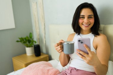 Evden uzakta çalışırken cep telefonu kullanan ve yatakta kahve içen mutlu serbest çalışan kadın.