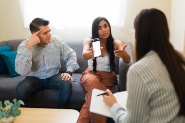 Latin Amerikalı çift profesyonel terapistle ilişki sorunlarını tartışıyor ve telefonda mesajlar gösteriyor.