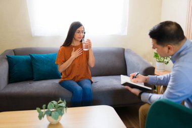 Terapi seansı sırasında psikoterapistle sorunlarını konuşurken su içen üzgün bir kadın.