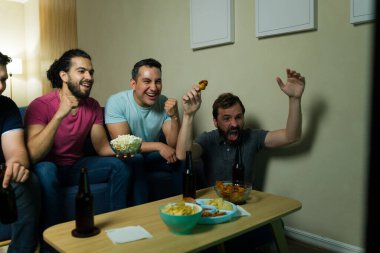 Bir grup erkek arkadaş, televizyonda maç izlerken tezahürat yapıyor ve kutlama yapıyorlar. Atıştırmalık ve içeceklerin tadını çıkarıyorlar.