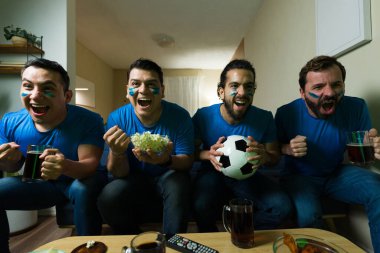 Geniş açılı, boyalı yüzlü dört İspanyol adamın evde televizyonda futbol maçı seyrederken tezahürat yapıp bira içmesi.