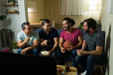 Latin erkek arkadaşlar televizyonda basketbol oynuyorlar, kanepede dinleniyorlar, bira içiyorlar ve sıcak bir evde atıştırmalık yiyorlar.