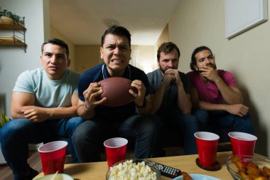 Bir grup erkek arkadaşın geniş açı görüntüsü gergin bir şekilde televizyonda futbol maçı izliyor, patlamış mısır yiyor ve evde bira içiyorlar.
