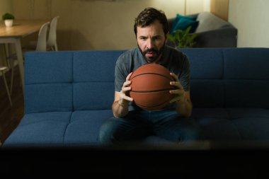 Adam gergin bir şekilde elinde top tutarken televizyonda basketbol maçı izliyor, evinde takımı için tezahürat yapıyor.