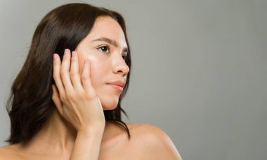 Güzel genç bir kadın nazikçe yüzüne dokunuyor, kusursuz cildini gösteriyor ve cildini ve güzelliğini ön plana çıkarıyor.