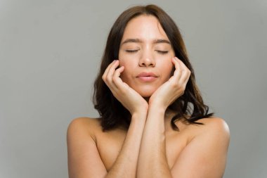 Gözleri kapalı İspanyol bir kadın elleriyle nazikçe yüzüne dokunuyor, cilt bakımını, sağlığını ve doğal güzelliğini teşvik ediyor.